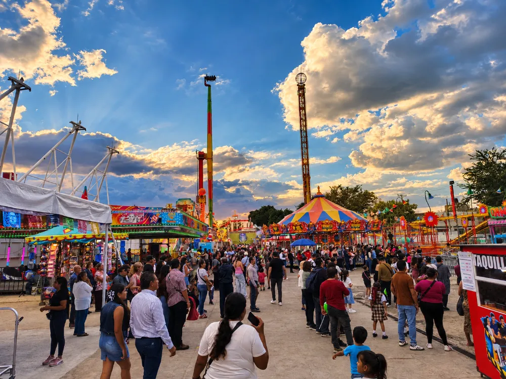 Familias recorren los juegos mecánicos de las Fiestas de Octubre en Guadalajara al atardecer, con carrusel y torres